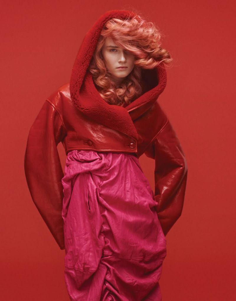 Fashion portrait of a female model with curly red hair wearing a red hooded jacket and pink skirt on a red background.