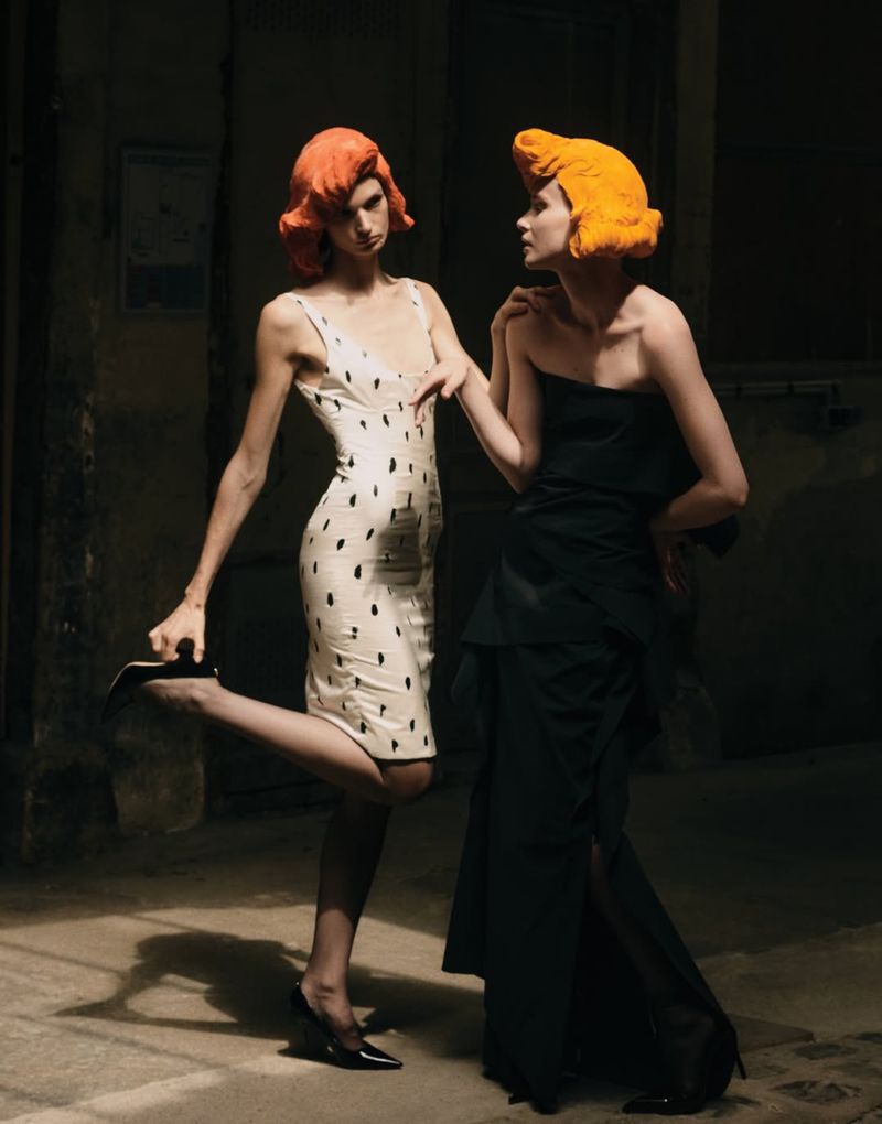 Two models with bright sculptural orange wigs wearing black and white dresses posing in dramatic lighting in an alley.
