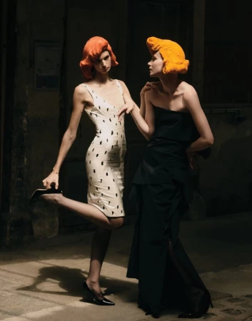 Two models with bright sculptural orange wigs wearing black and white dresses posing in dramatic lighting in an alley.