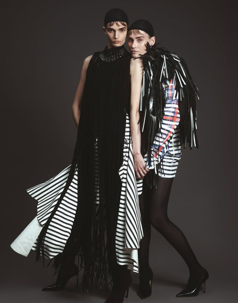 Two models in black and white striped dresses pose in a studio. One wears a long black fringe dress, the other a black leather-look shawl.