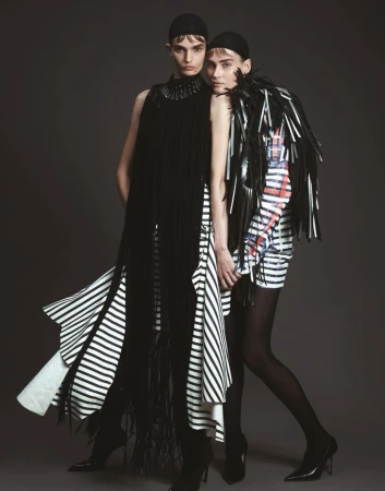 Two models in black and white striped dresses pose in a studio. One wears a long black fringe dress, the other a black leather-look shawl.
