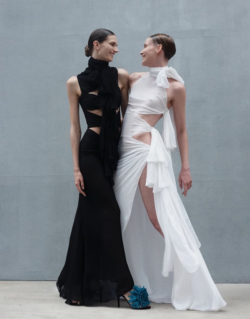Two women stand close together in a studio setting, showcasing elegant, modern dresses in black and white. They are smiling and looking at each other.