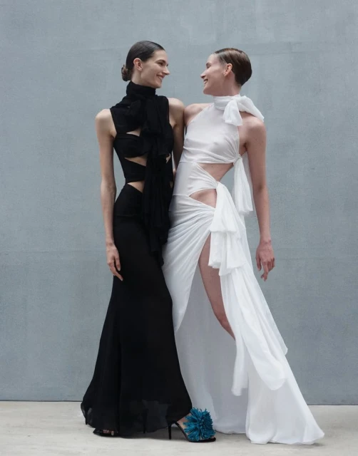 Two women stand close together in a studio setting, showcasing elegant, modern dresses in black and white. They are smiling and looking at each other.