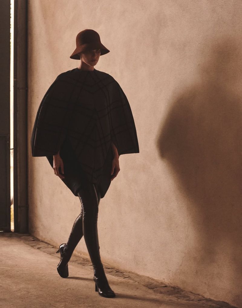 A model in a brown hat and cape walks in front of a tan wall with a shadow, showcasing fashion in an indoor setting with neutral tones.
