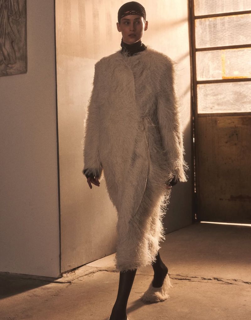 Woman in a fashionable, off-white, shaggy coat walks in a minimalist interior with concrete floors and a textured wall in the background.