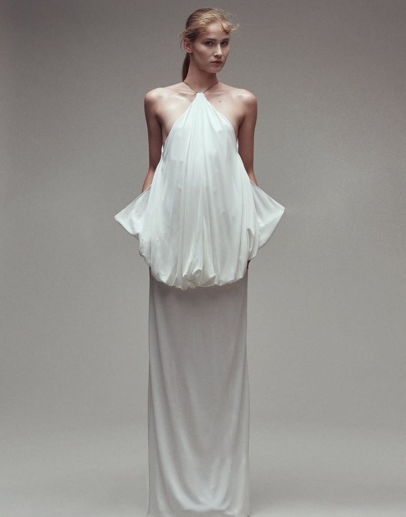 Model wearing a structured white halter dress standing against a neutral grey studio background for Vogue Adria