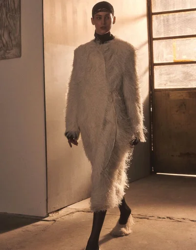 Model walking forward wearing a textured cream coat and dark cap in an interior space with warm light filtering through a glass door