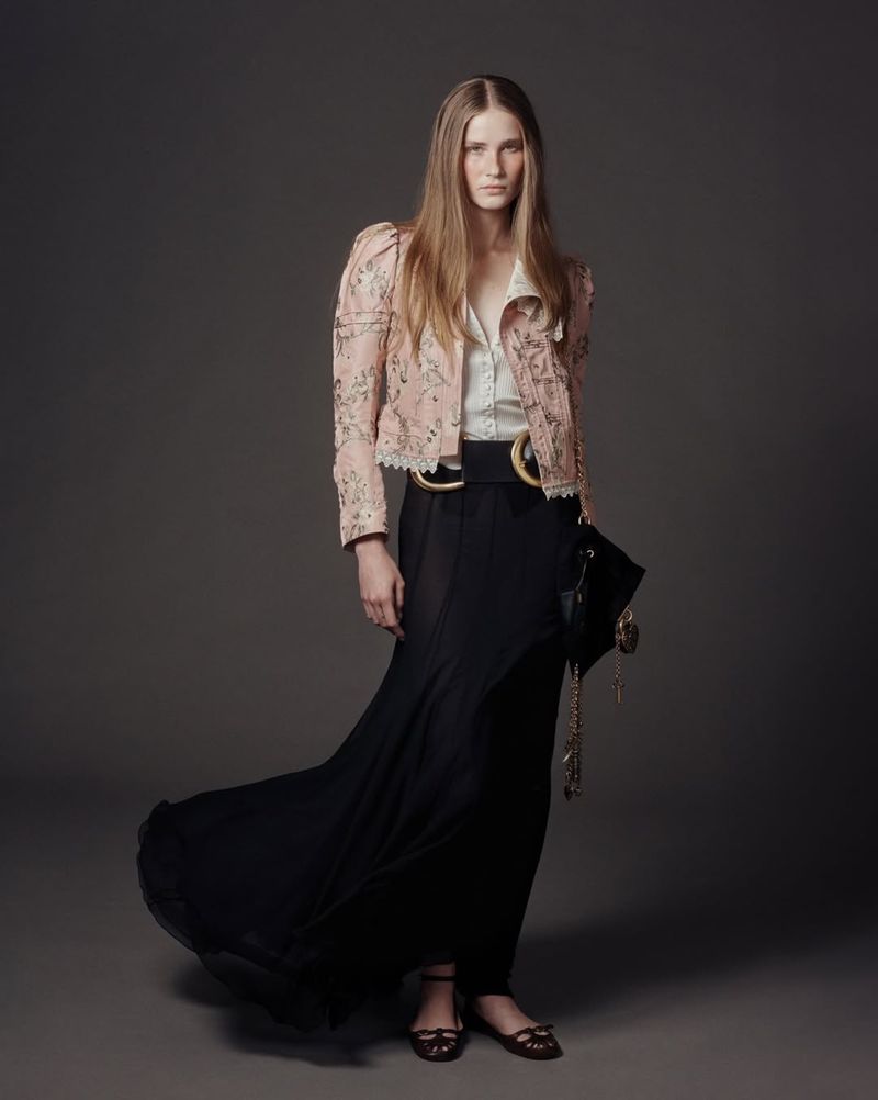 Model with long blonde hair wearing a patterned pink jacket and long black skirt against a grey background.