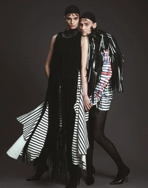 Two female models wearing black caps and avant-garde fringe apparel against a dark grey seamless studio backdrop for Vogue Adria.