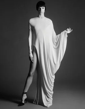 Model in a long white draped dress with high leg slit and dark heels against a grey studio backdrop.