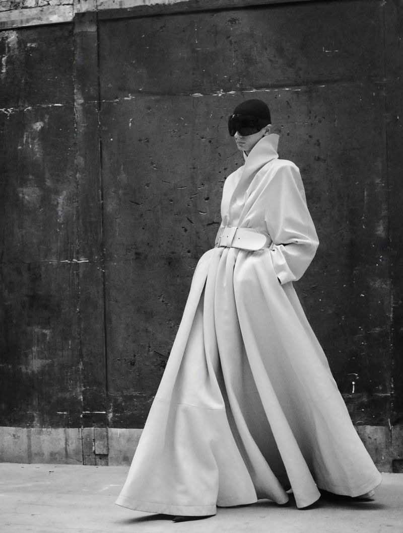 Model wearing a structured light-colored coat and dark sunglasses against a dark textured wall