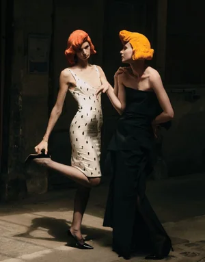 Two models with bright sculptural orange wigs wearing black and white dresses posing in dramatic lighting in an alley.