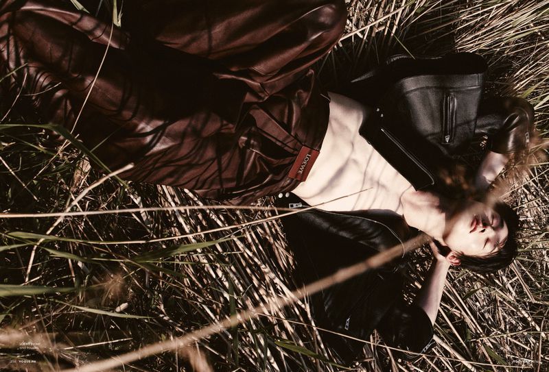 Fashion shot of a supine person wearing a black leather jacket and maroon leather pants, lying in tall grass in an outdoor setting.