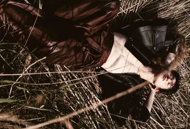 Fashion shot of a supine person wearing a black leather jacket and maroon leather pants, lying in tall grass in an outdoor setting.