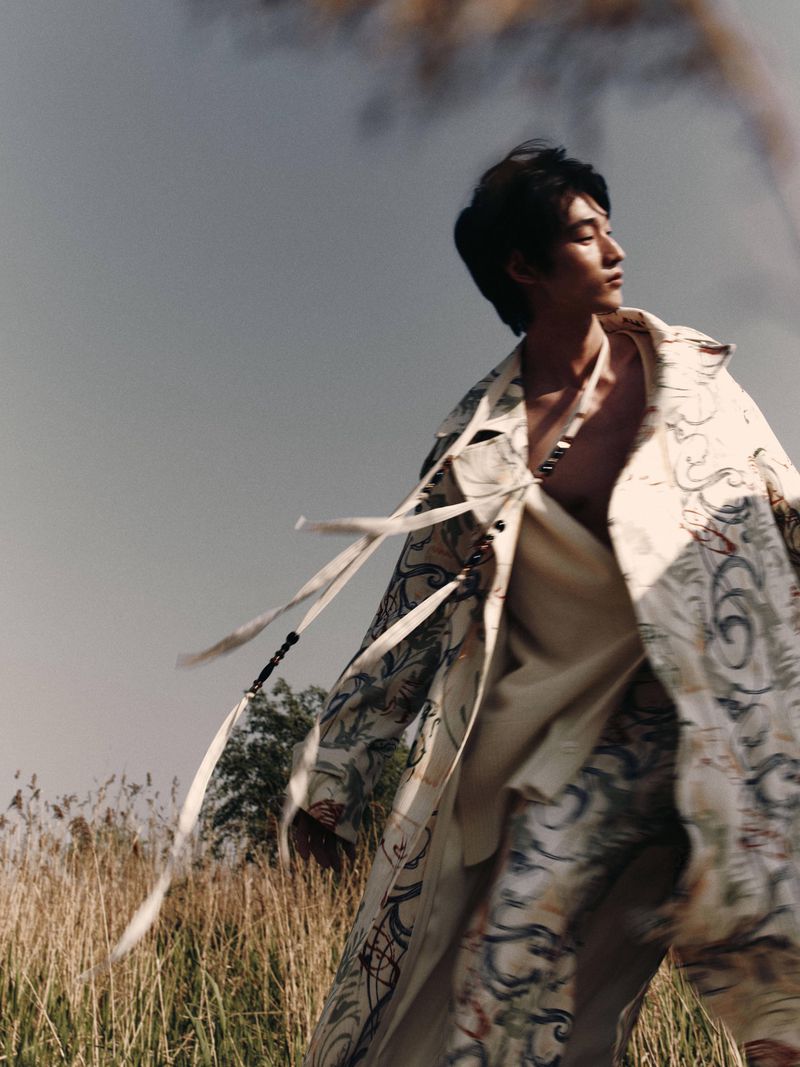 Asian male model in patterned coat and pants poses outdoors in a field of tall grass under a cloudy sky in a fashion editorial style.