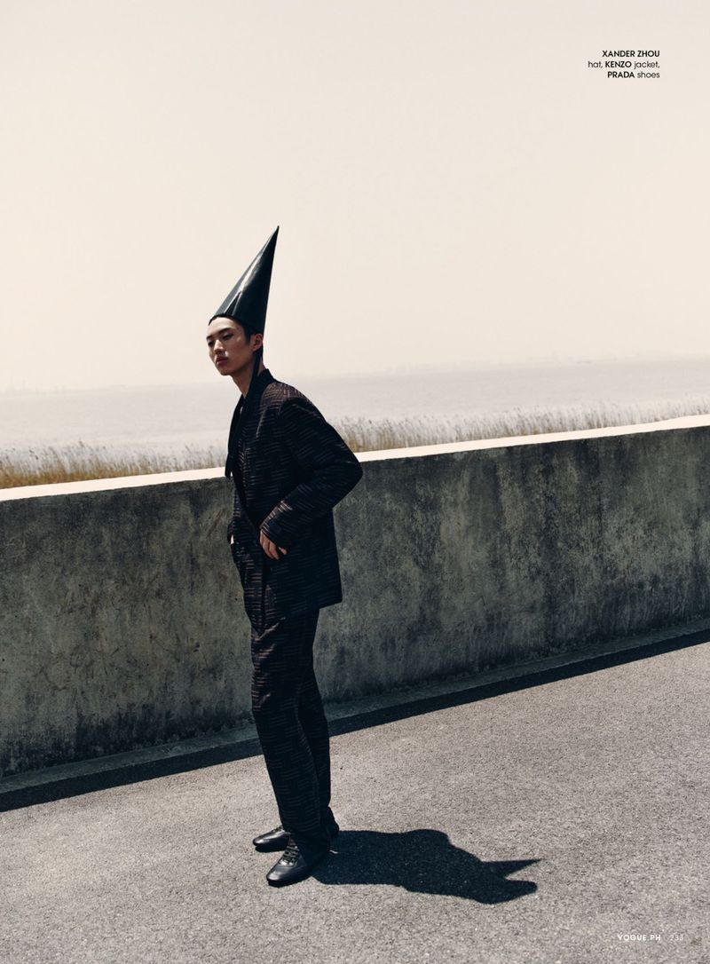 An Asian male model poses in a textured black suit and a shiny black cone hat on an overcast day in an outdoor setting, fashion editorial.