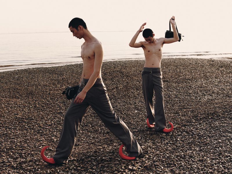 Two shirtless men with trousers and unique shoes stand on a rocky beach at sunset, one holding a bag, looking towards the sea.