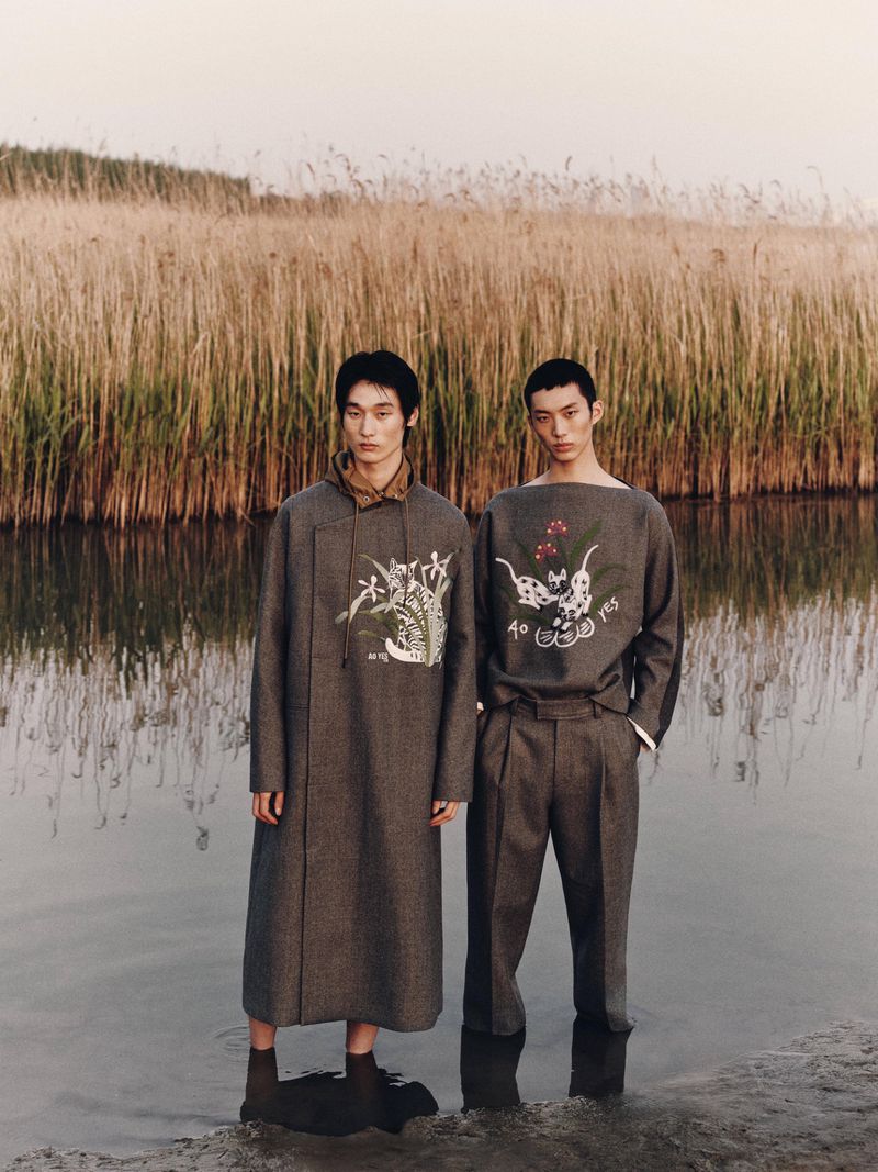 Two male models in stylish, grey patterned outfits stand in shallow water, set against tall reeds and a light sky backdrop.