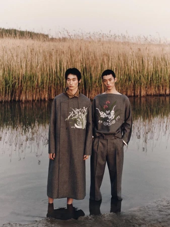 Two male models in stylish, grey patterned outfits stand in shallow water, set against tall reeds and a light sky backdrop.