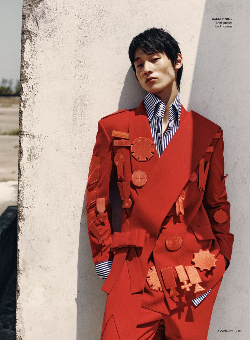 Asian male model in a bright red, heavily decorated suit leans against a weathered wall in an outdoor editorial fashion shot.
