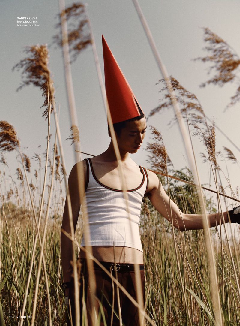 Young adult male in tank top stands in tall grass, wearing a red cone hat in an outdoor fashion editorial shot under a blue sky.