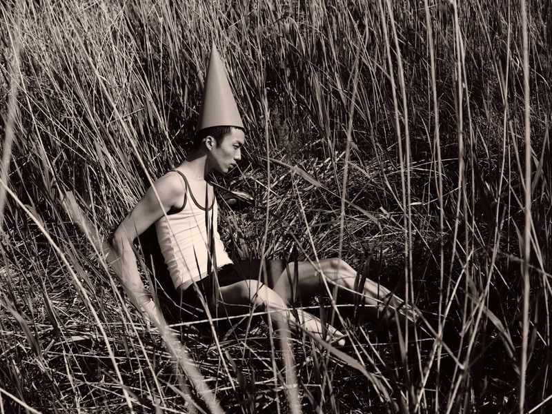 An Asian man wearing a cone hat sits among tall grasses in a black and white outdoor photograph taken at eye-level.