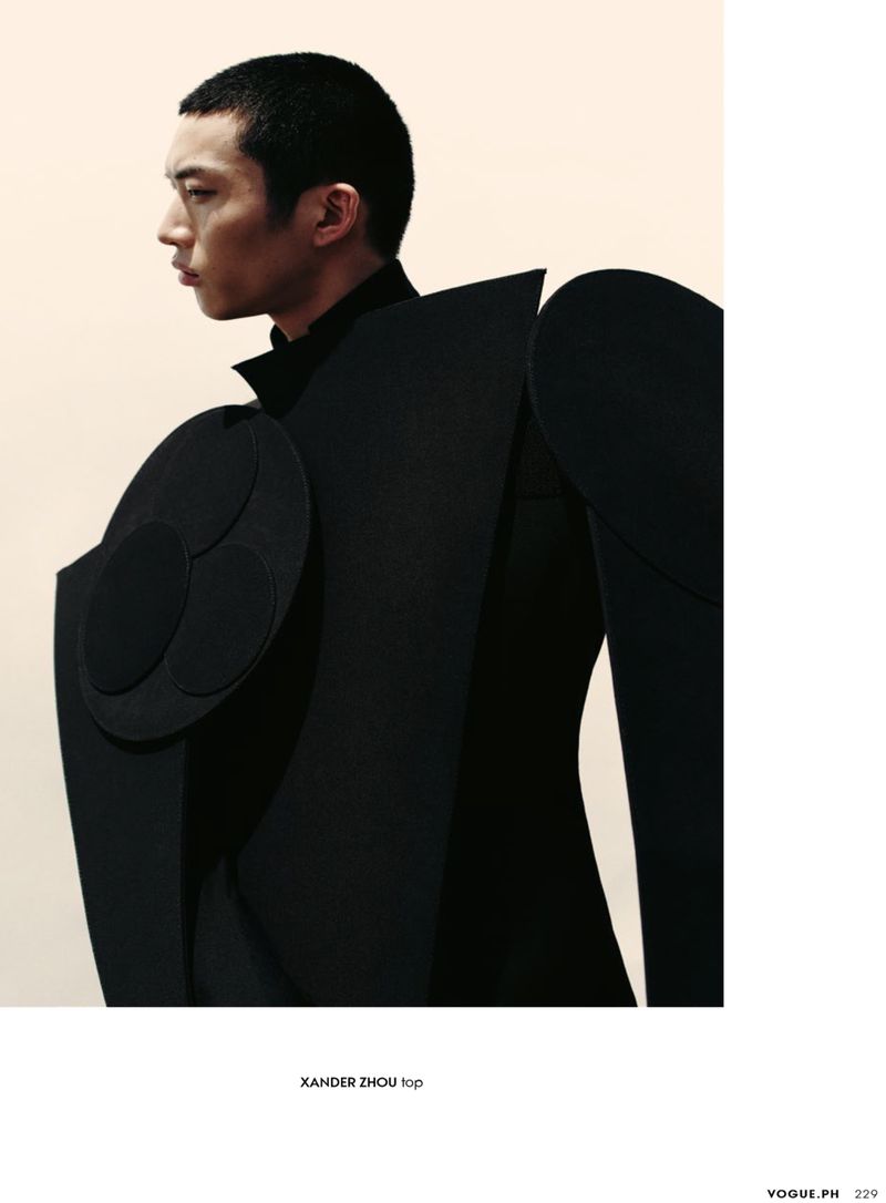Profile shot of an Asian male model wearing an avant-garde, oversized black garment with circular shoulder detail against a beige backdrop.