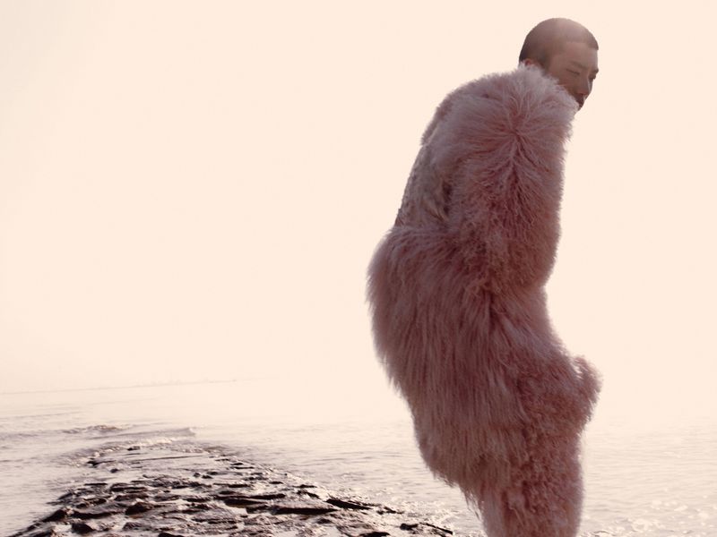 Adult wearing a shaggy pink coat standing on a rocky beach in front of a misty body of water on an overcast day.