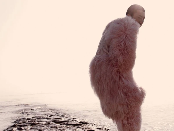 Adult wearing a shaggy pink coat standing on a rocky beach in front of a misty body of water on an overcast day.