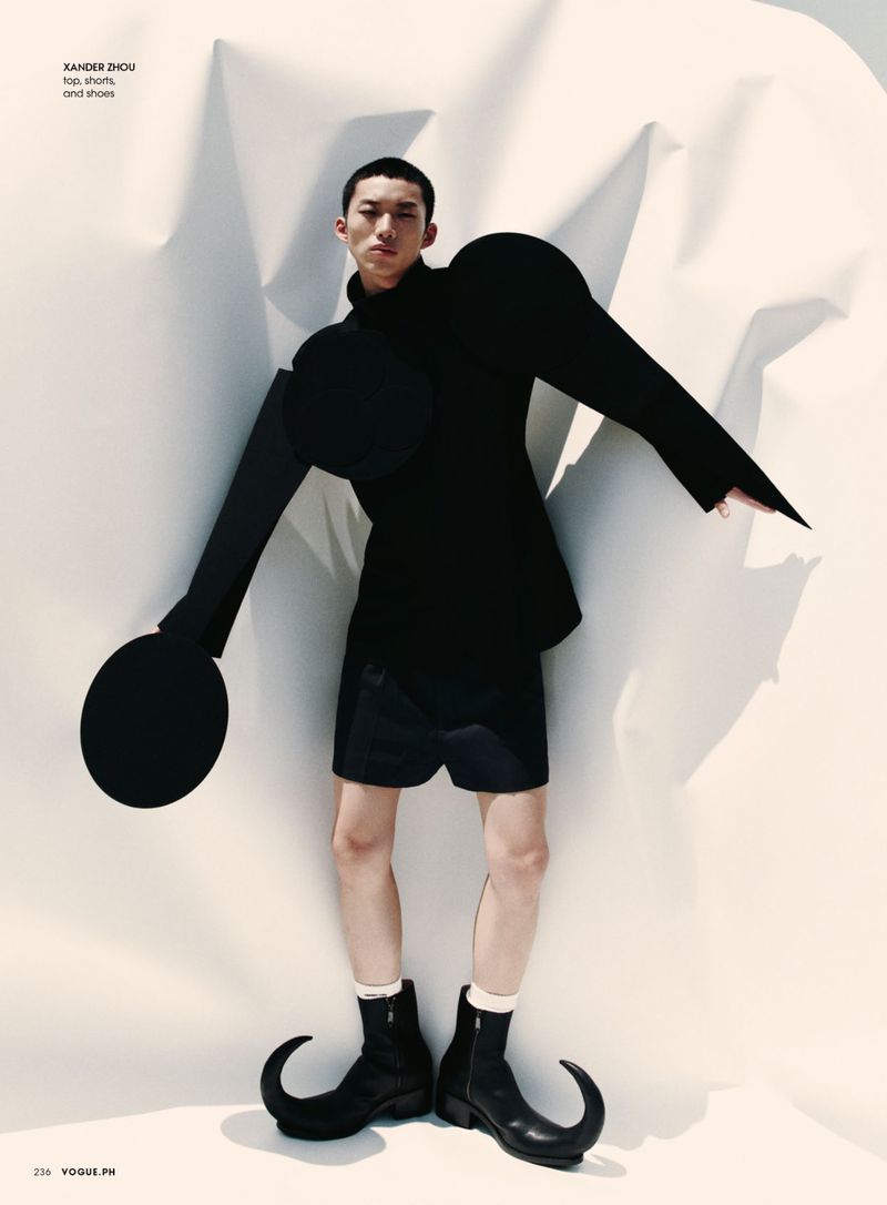 Editorial fashion shot of an Asian male model wearing a black jacket and shorts with sculptural paper art background and fantasy-style boots.