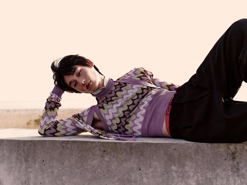 Male fashion model poses in patterned top and dark pants, reclining on a concrete ledge against a light, natural backdrop.