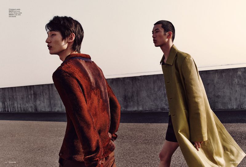 Two Asian men in fashionable outerwear walk on a rooftop with minimalist style, showcasing contemporary menswear in a neutral-toned setting.