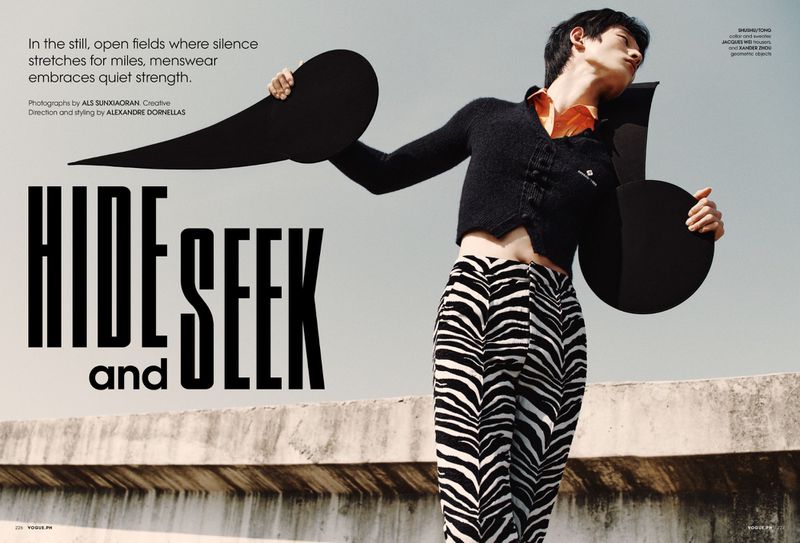 Fashion editorial shot of an Asian male model in black cardigan and zebra-striped pants, holding black geometric shapes outdoors.