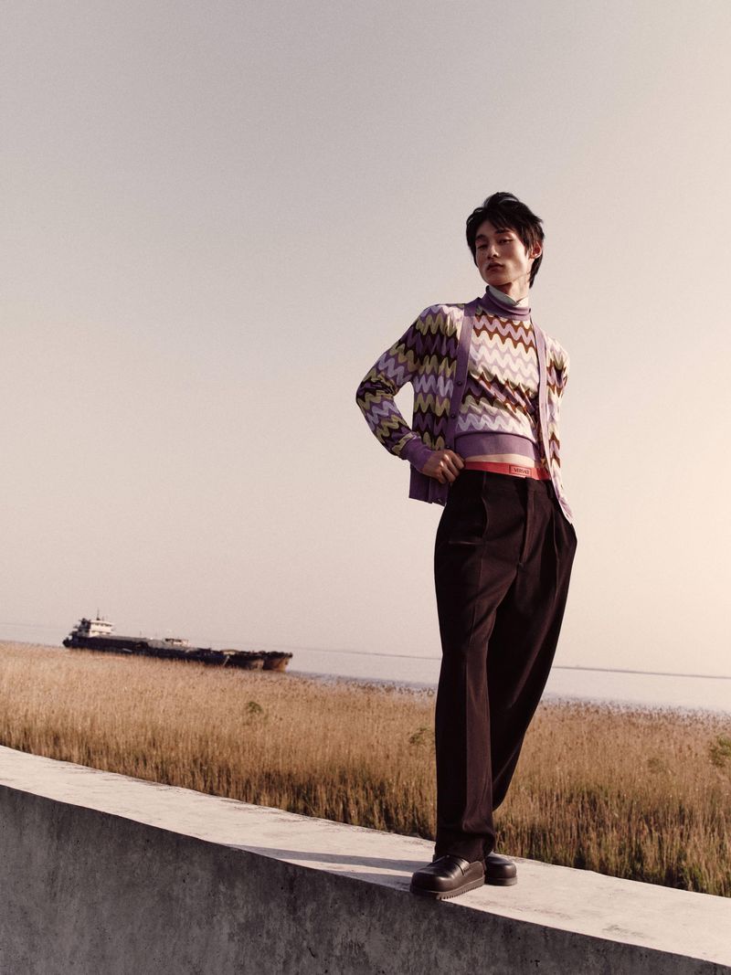 A stylish young man poses outdoors wearing patterned sweater, cardigan, and dark trousers, with a distant ship on the water.