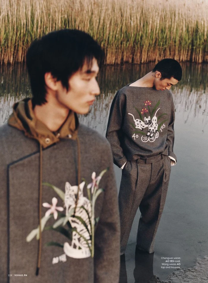 Two male models in grey embroidered tops standing near a shallow river bank with tall reeds in the background