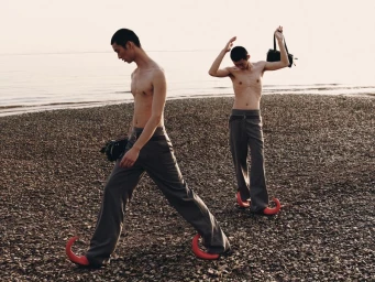 Two shirtless male models wearing grey trousers and curved red shoes on a rocky beach.