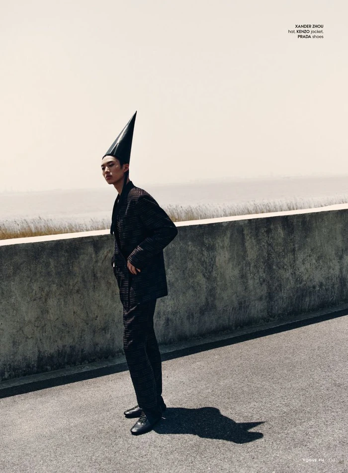 Model wearing a dark suit and tall pointed hat standing on an asphalt road near a concrete wall.