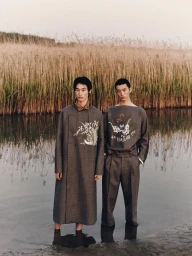 Two male models standing in shallow water before a tall reed-covered bank wearing grey embroidered garments