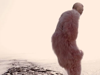 Model wearing a textured pink furry coat standing near a rocky shoreline by the ocean