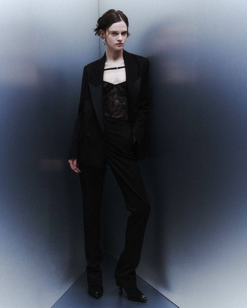 A fashionable adult female poses indoors wearing a black blazer, lace top, and pants, standing in front of a gradient wall in a studio shot.