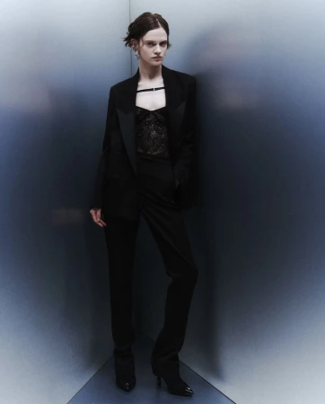 A fashionable adult female poses indoors wearing a black blazer, lace top, and pants, standing in front of a gradient wall in a studio shot.
