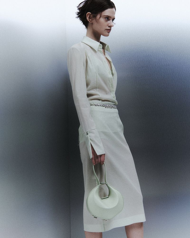 A woman in a light green shirt and skirt holding a round handbag in a studio with a gray background in a fashion shot.