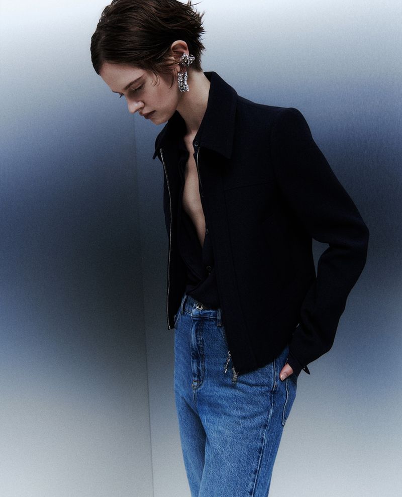 A young adult woman poses in a dark zip jacket and blue jeans against a blue gradient background, showcasing fashion style.