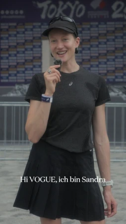 Smiling woman in black athletic wear stands in front of Tokyo Olympics banner, wearing a cap, holding a microphone and looking at the camera.