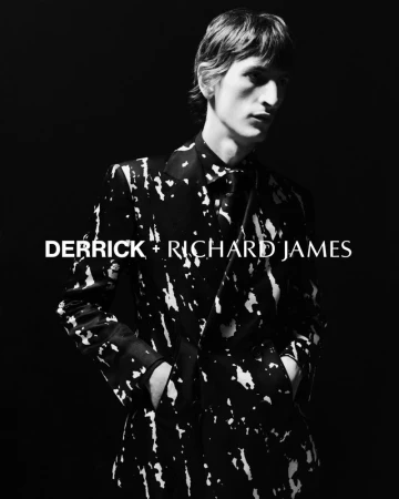 Monochrome fashion portrait: a slender male model in a patterned suit stands against a dark background, showcasing modern menswear.