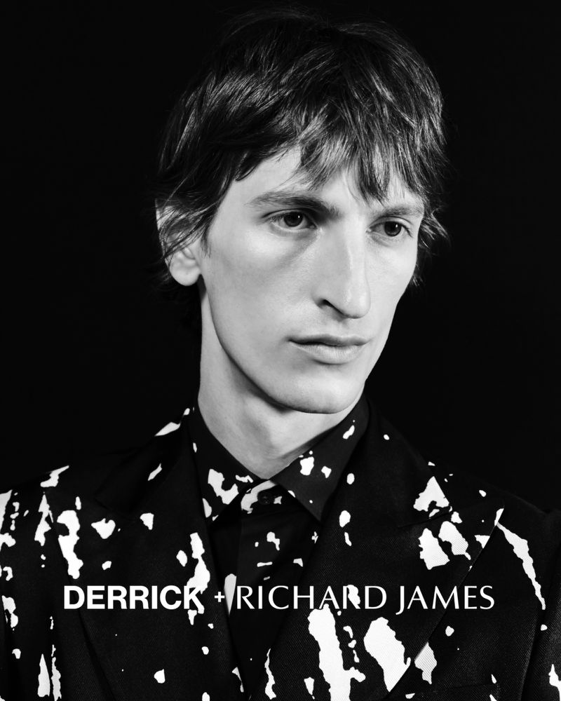 Black and white portrait of a fair-skinned adult male with dark hair wearing patterned suit and shirt against a solid black background.