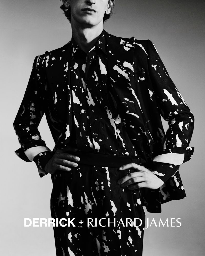 Model Theodor Pal wearing a black and white splash-print tailored suit with hands on hips against a studio backdrop.