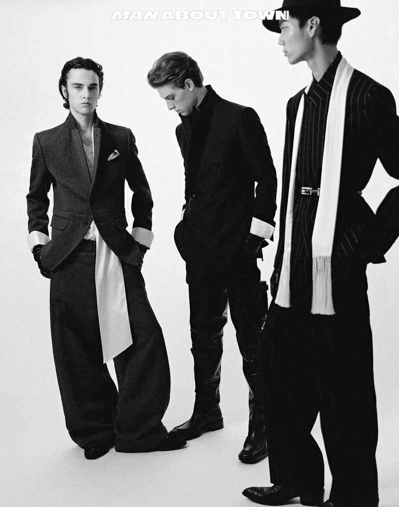 Three fashionable men in black suits pose in a photo studio with a white backdrop. The monochrome image showcases runway-ready fashion.