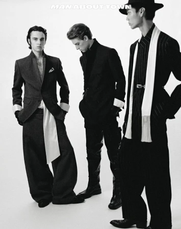 Three fashionable men in black suits pose in a photo studio with a white backdrop. The monochrome image showcases runway-ready fashion.