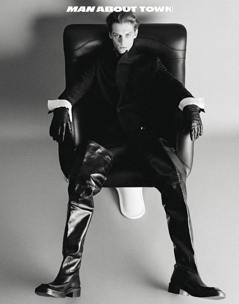 Monochrome portrait of a slender male model in high leather boots, seated on a stylized toilet seat in a photo studio.